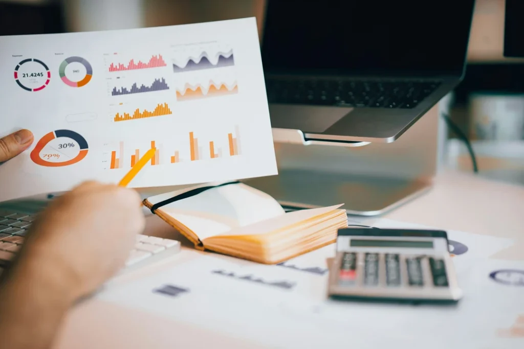 Pessoa a analisar gráficos e indicadores financeiros num escritório com calculadora e computador portátil.